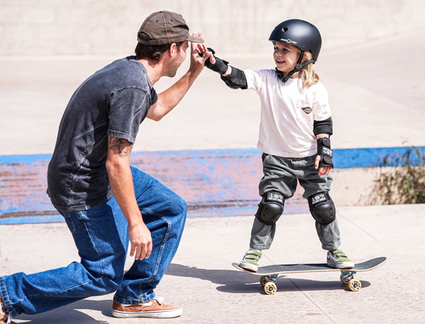 Man and child at a skate park with the man giving a high-five to the child on a skateboard wearing 187 Killer Pads.